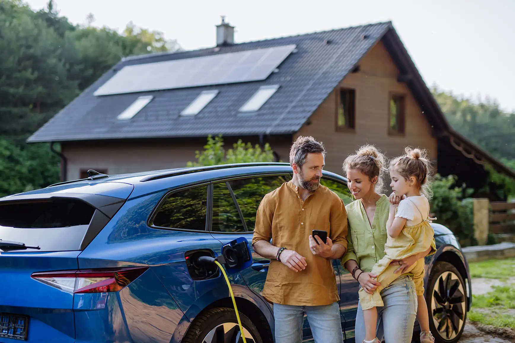 Junge Familie steht vor einem Elektroauto und im Hintergrund ist ihr Eigenheim mit einer Photovoltaikanlage zu sehen.